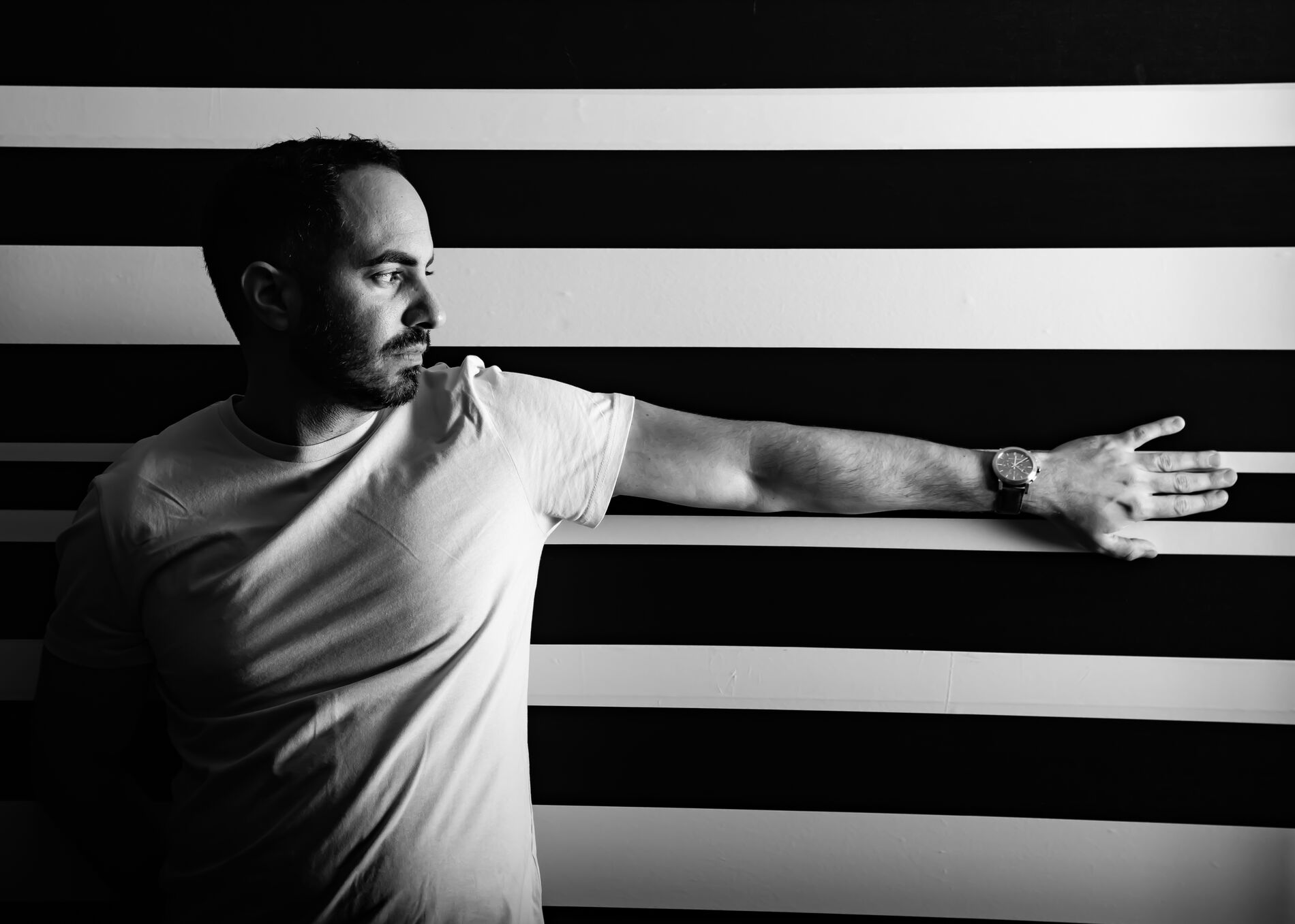 Black and white photo of SOHEILL in profile, arm extended along a striped wall.