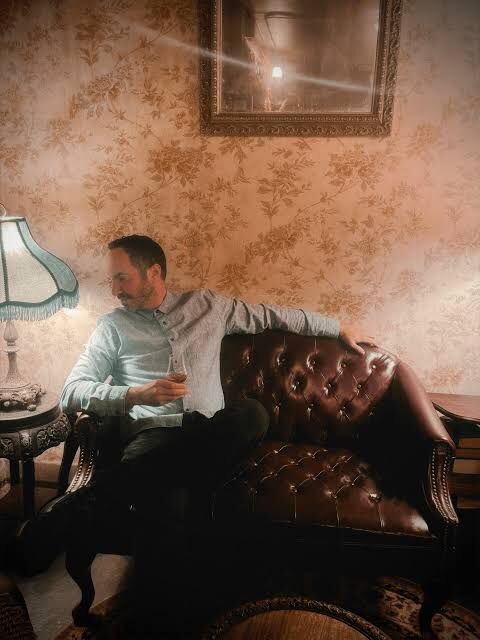 SOHEILL relaxed on a vintage leather couch, glass in hand, warm lamp light.
