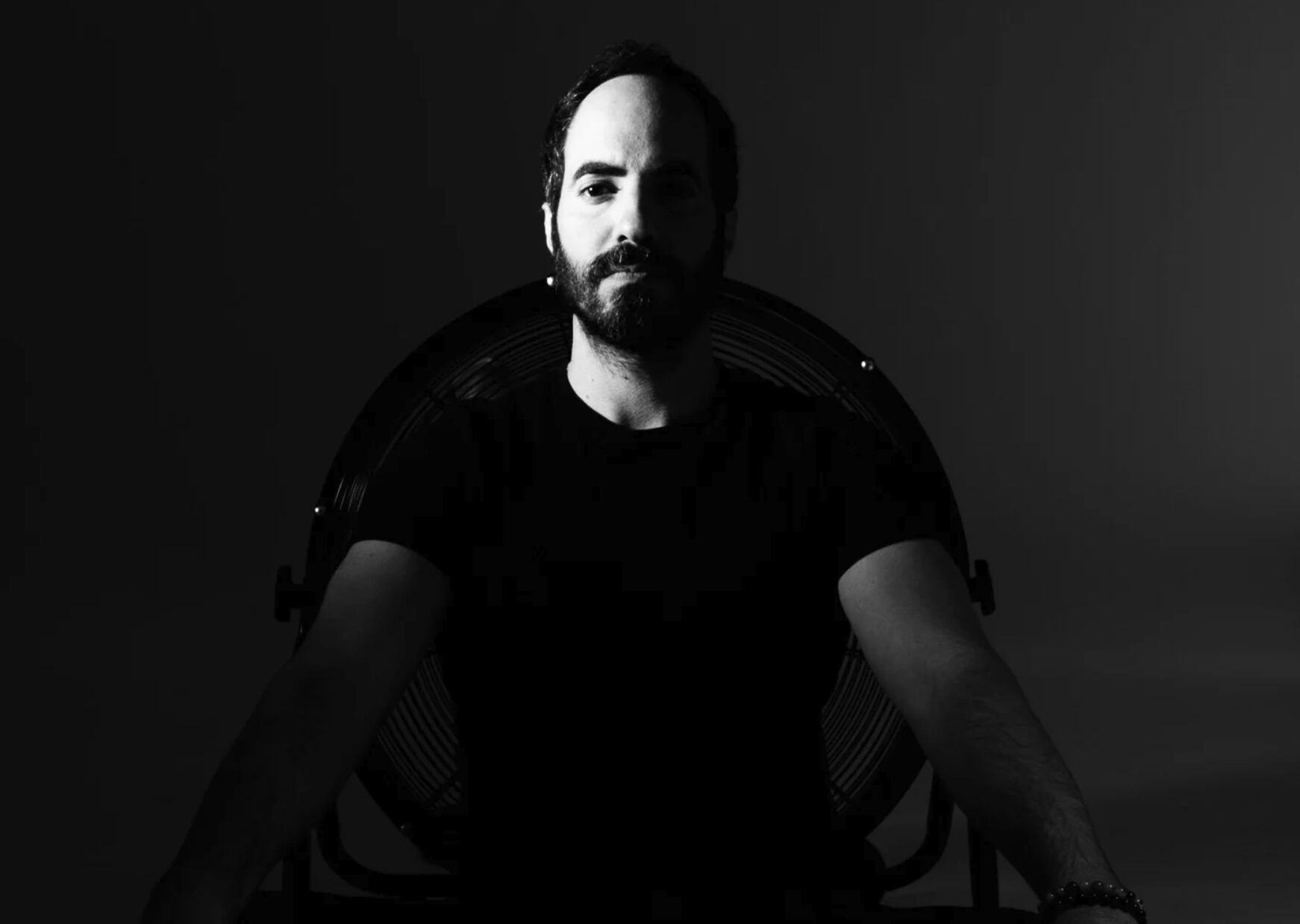 SOHEILL standing in near-total darkness, arms outstretched, faint studio light behind him.