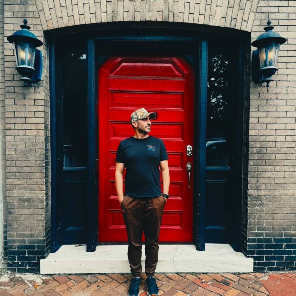 SOHEILL standing before a red door, camo cap, looking off to the side.