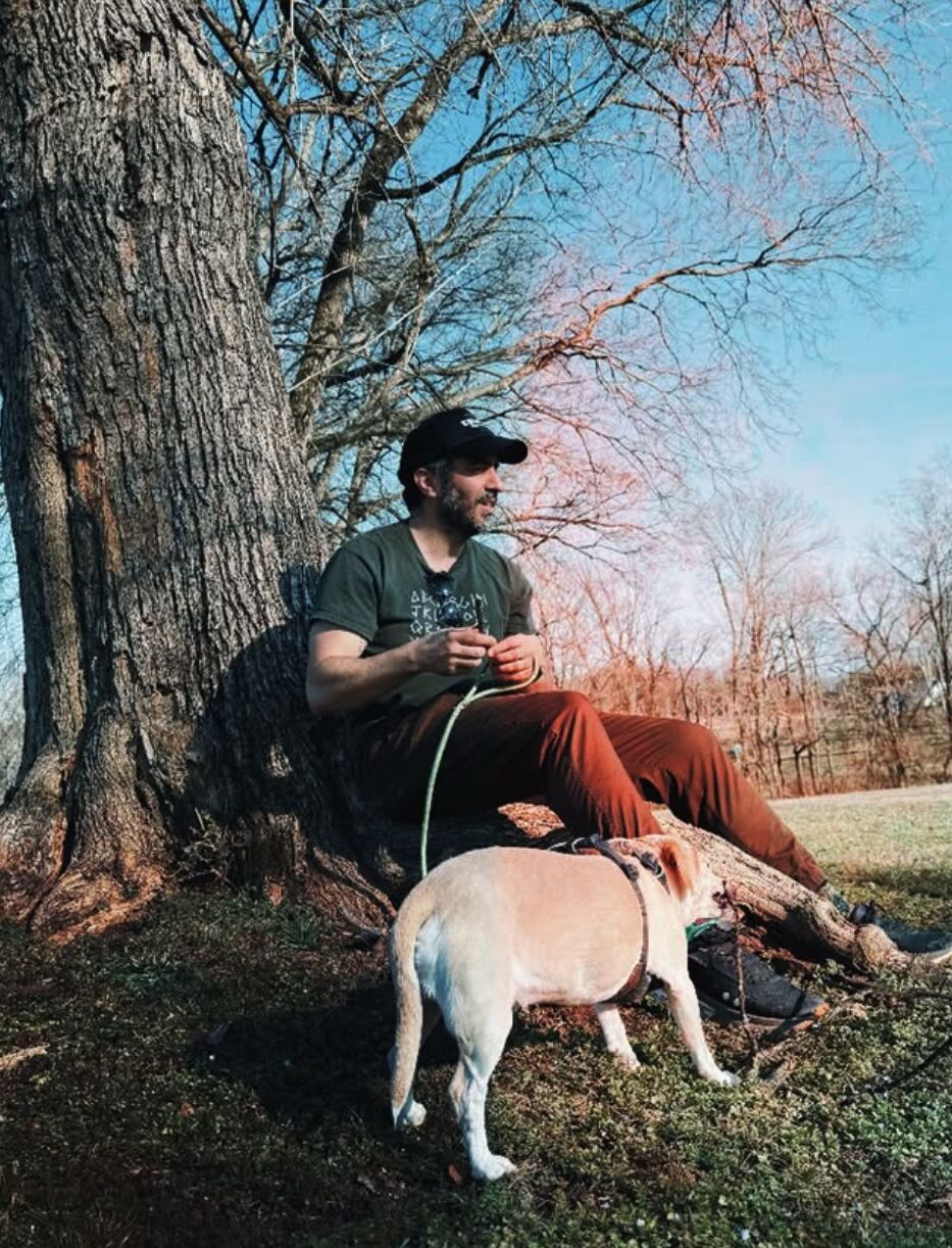 SOHEILL sitting against a tree outdoors with his dog on a leash.