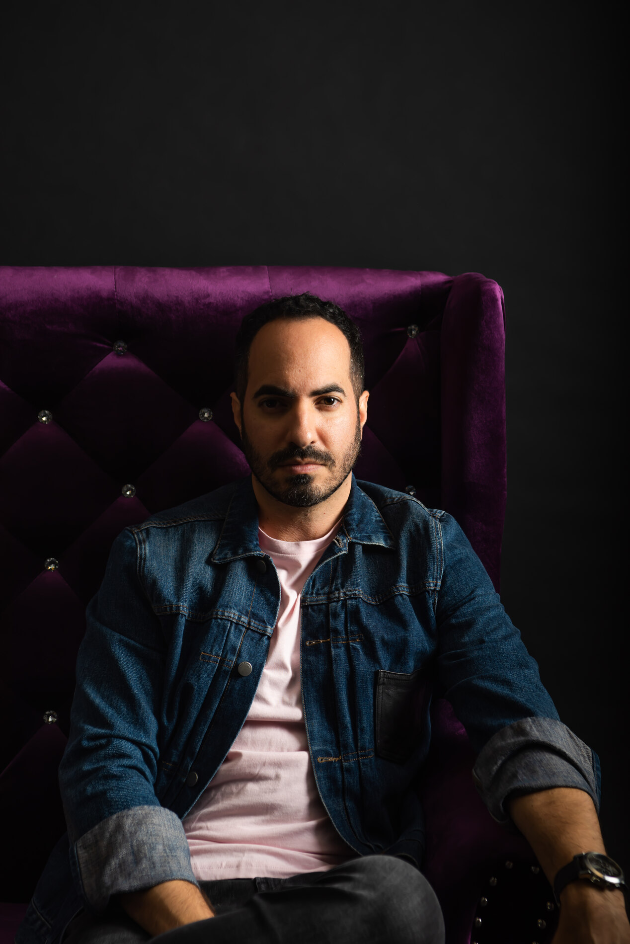 SOHEILL seated in a purple velvet chair, denim jacket over a pink tee, direct gaze.